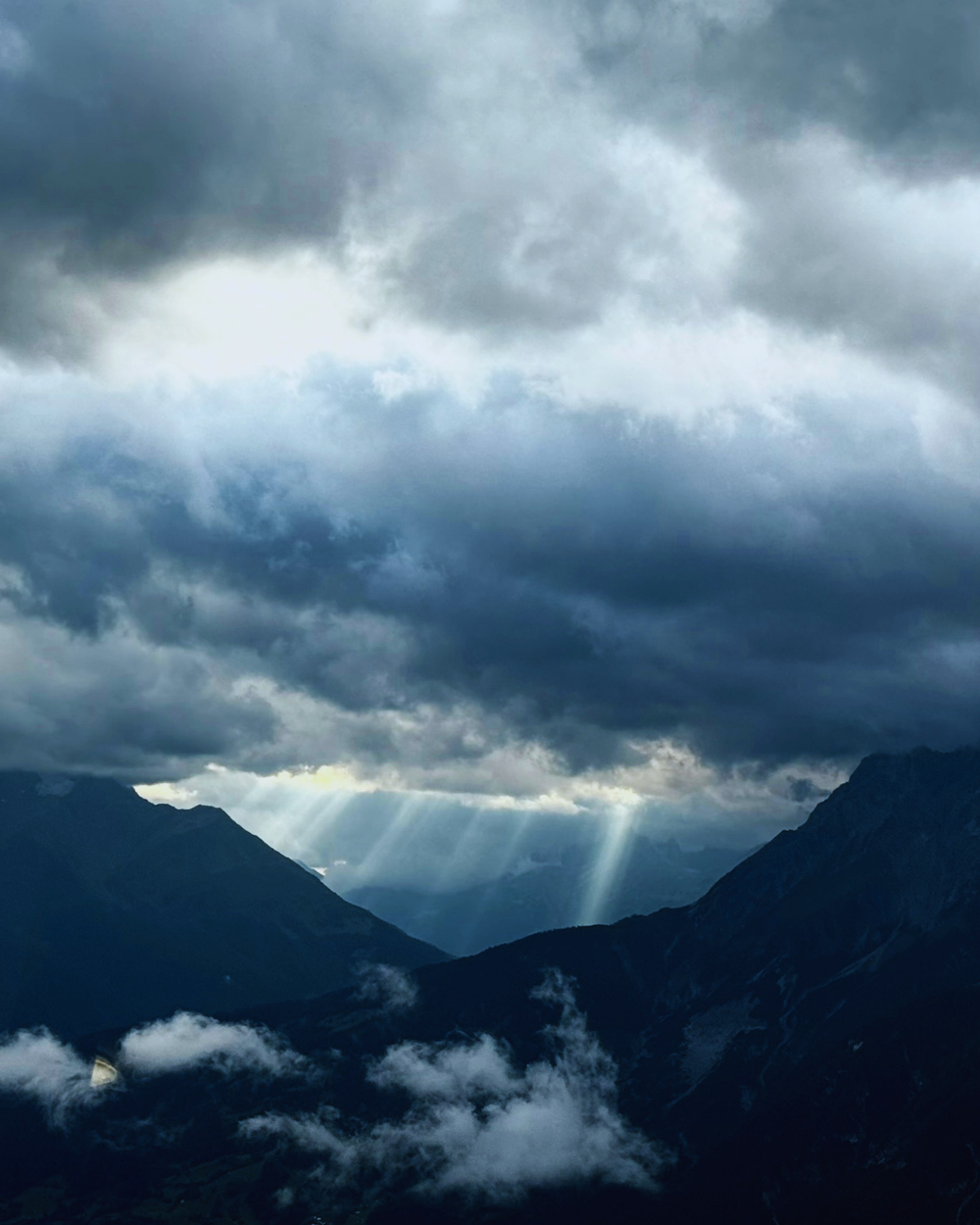 Dunkle Gewitterwolken über einem Bergtal, durch eine Wolkenlücke fallen Sonnenstrahlen zwischen die Bergflanken